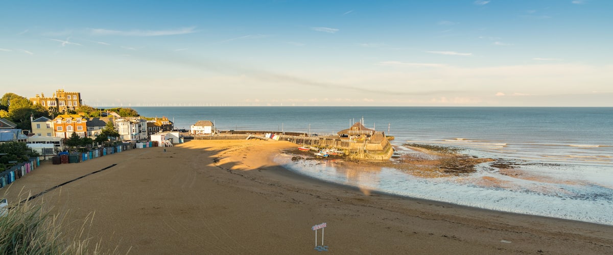 A coastal beach with a rocky outcrop, a pier, and colorful buildings in the background.
