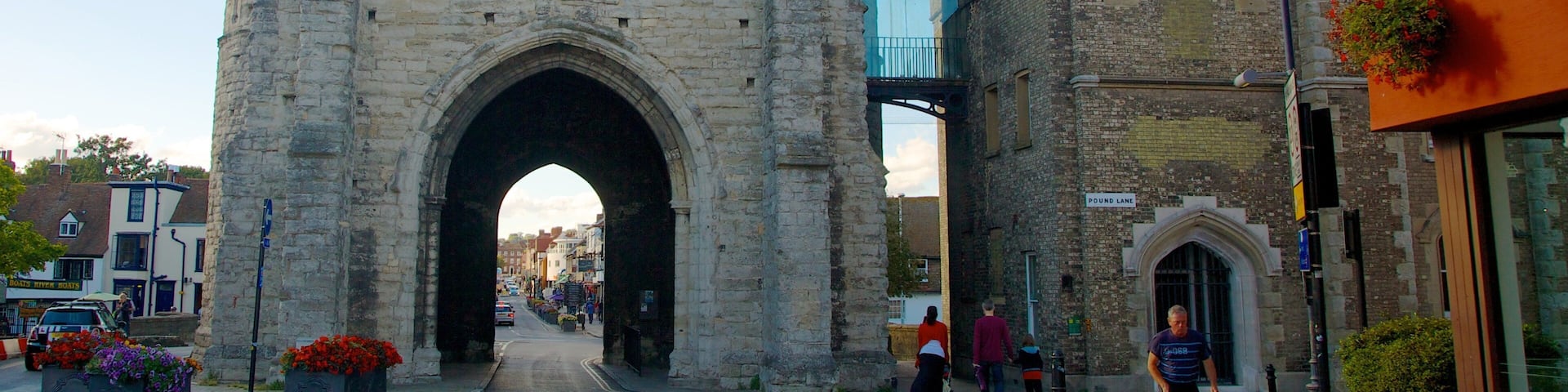 A historic stone archway with a tower, surrounded by modern buildings and a street with pedestrians.