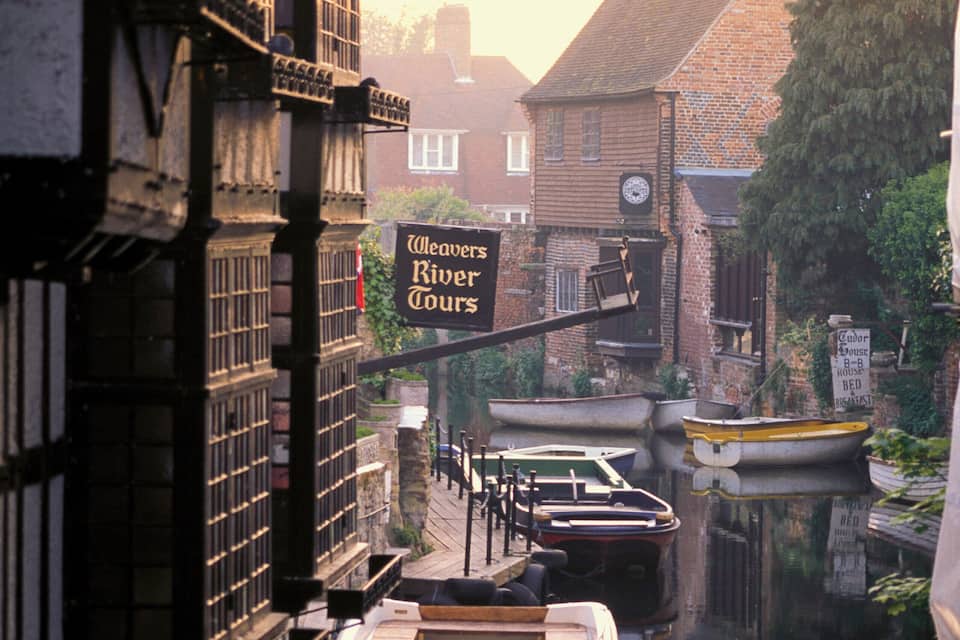 A narrow canal with boats docked along the sides, a building with a sign for Weavers River Tours, and a clock tower in the background.