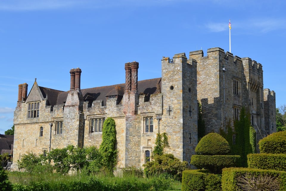 A historic stone castle with multiple chimneys, a flag on top, and well-maintained hedges.