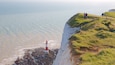 A coastal landscape with a lighthouse on a rocky shore, grassy cliffs, and people standing on the cliff edge.