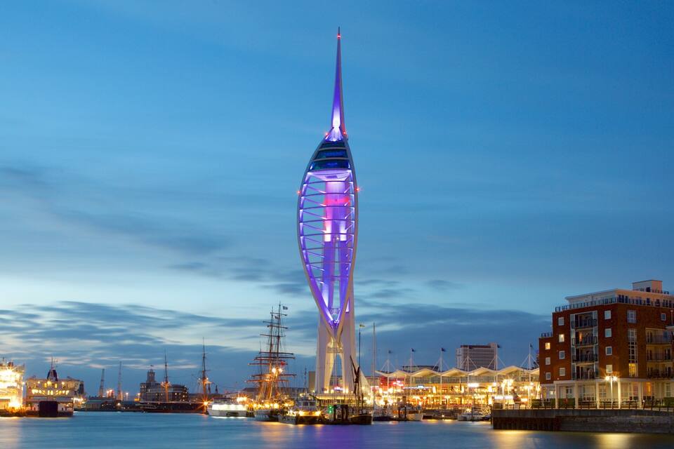 A modern tower with a spire, surrounded by a marina with boats and buildings.