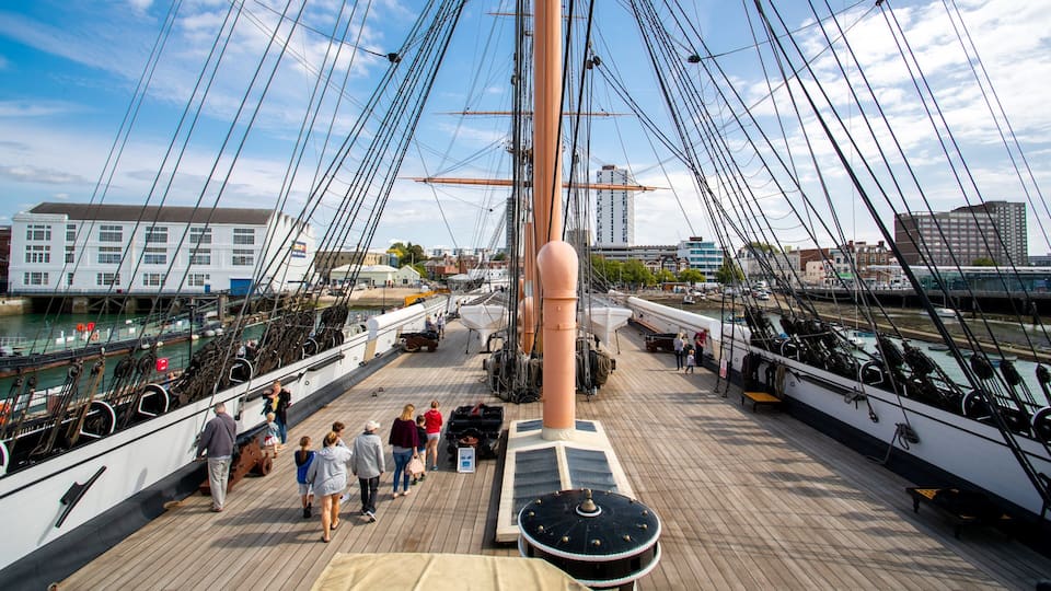 A wooden deck with people walking, a large ship's mast, and buildings in the background.