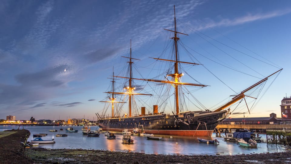 A large sailing ship docked at a harbor with several smaller boats around it, under a twilight sky.