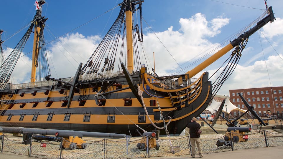 A large, yellow and black ship with multiple masts and rigging, docked at a port with a building in the background.