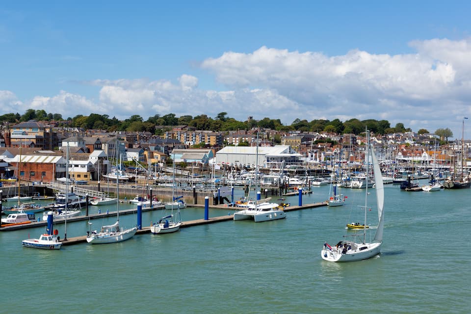 A marina filled with boats, a sailboat in the foreground, and a town with buildings in the background.