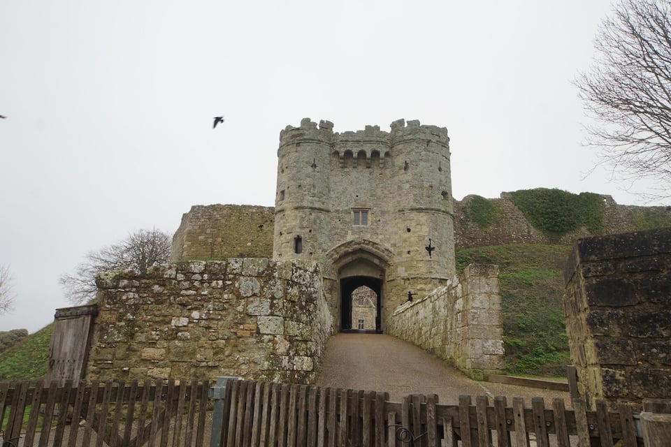 Un castillo medieval con una torre central, una valla de madera y un sendero que lleva a la entrada.