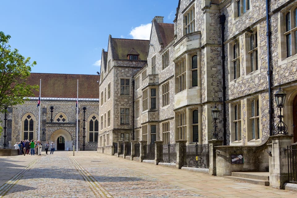 A cobblestone street lined with historic stone buildings, a flagpole, and a group of people.