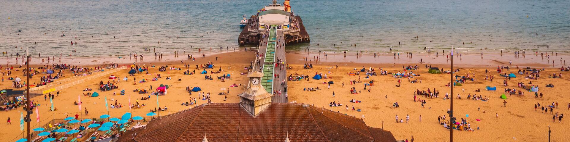 A beach with a pier extending into the sea, surrounded by people and umbrellas.
