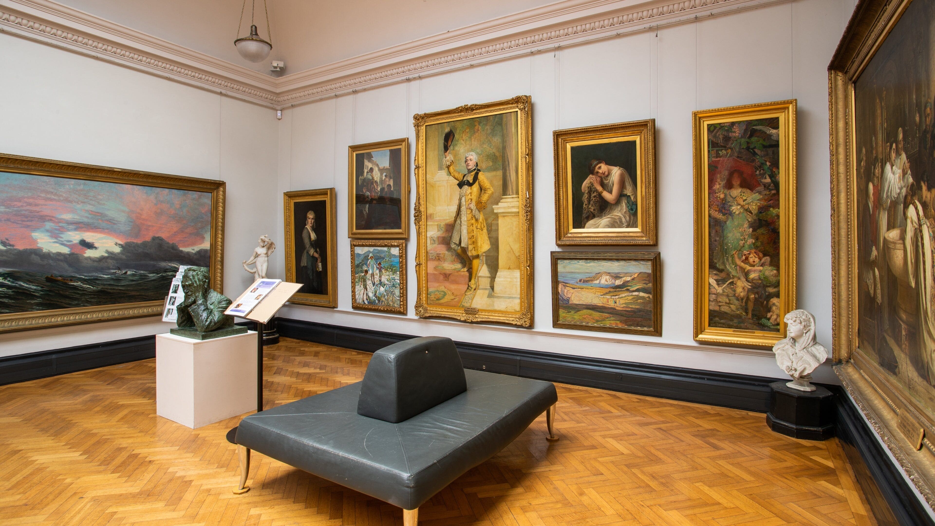 A museum interior with classical paintings and sculptures on the walls, a black leather couch, and a wooden parquet floor.