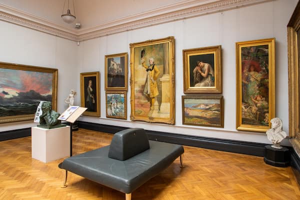 A museum interior with classical paintings and sculptures on the walls, a black leather couch, and a wooden parquet floor.