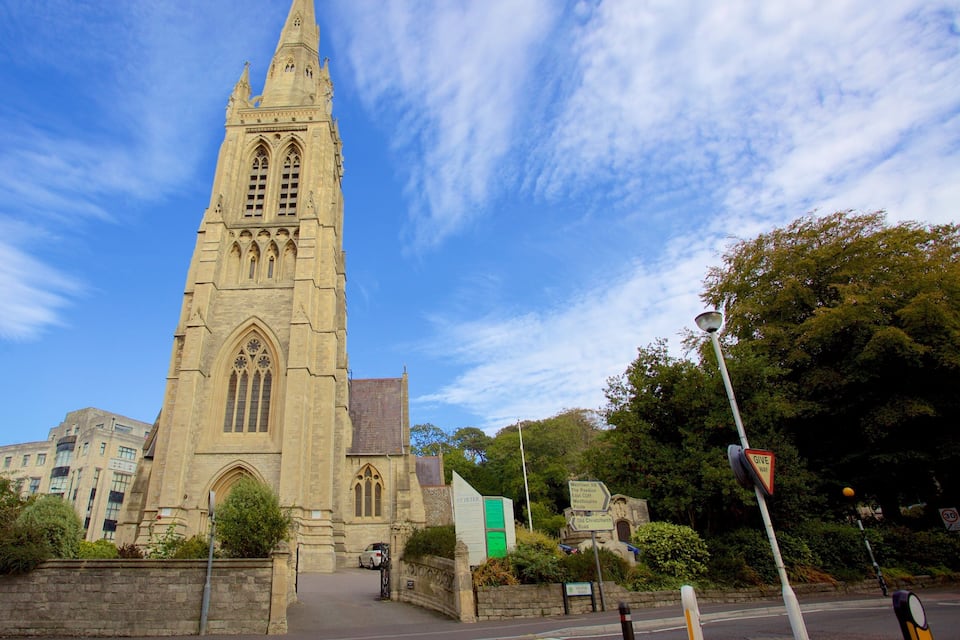 A tall church with a pointed spire, surrounded by trees and modern buildings.