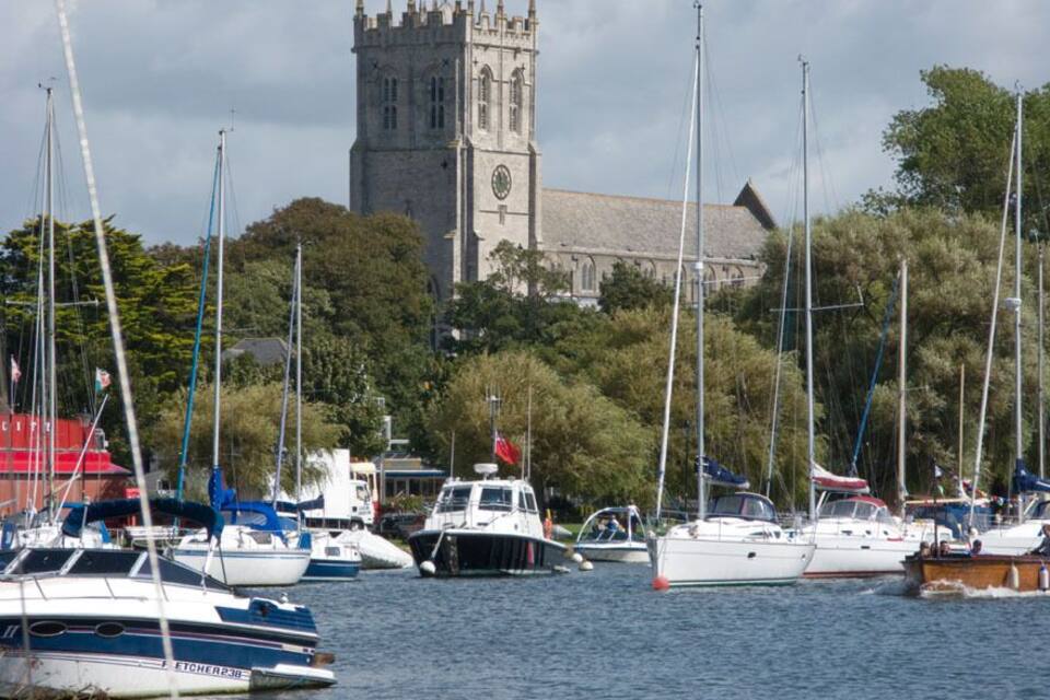 A marina with several sailboats docked, a church with a clock tower, and lush greenery in the background.