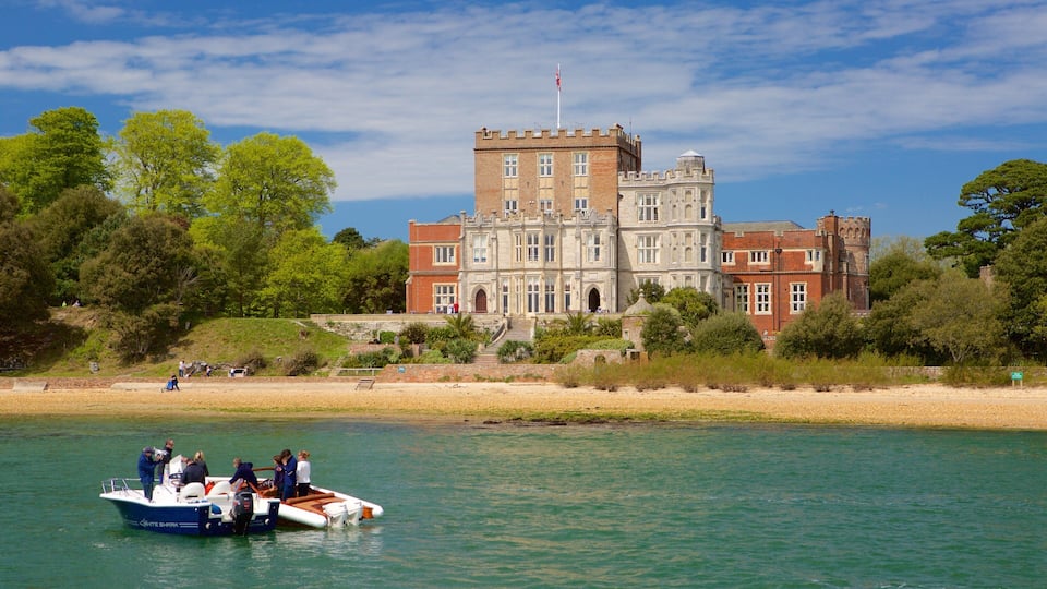 A historic building with a castle-like tower, a boat on the water, and a beach with people and greenery.