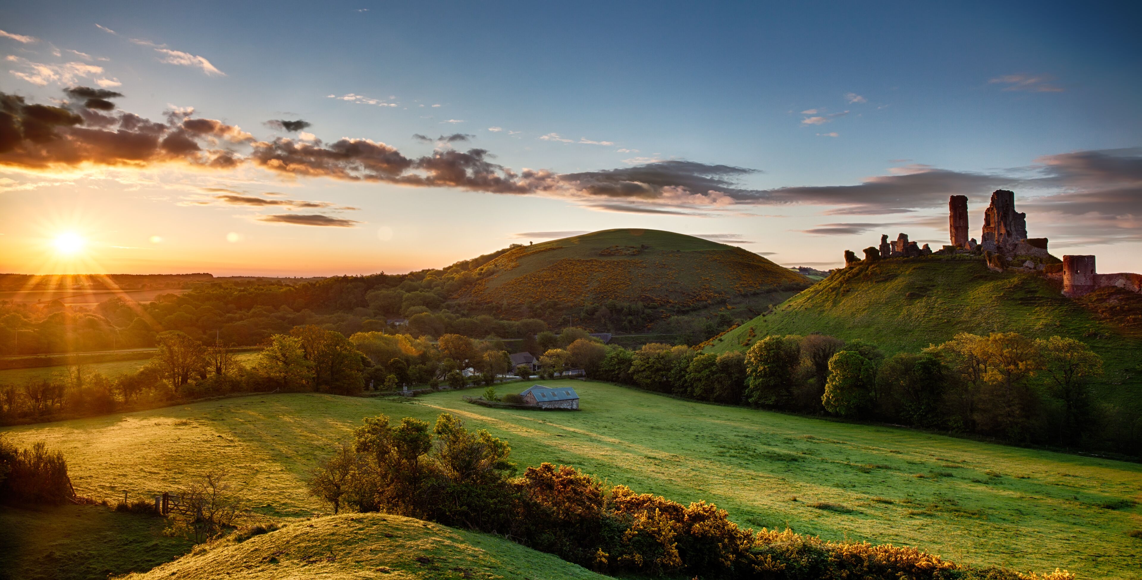 A sunset over a grassy hill with ruins and a small house.