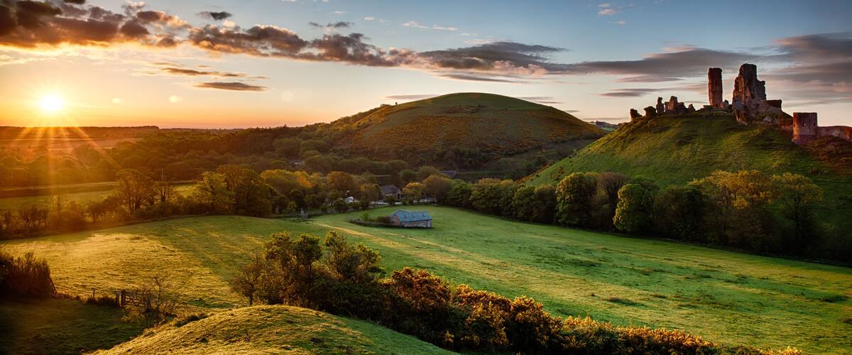 A sunset over a grassy hill with ruins and a small house.