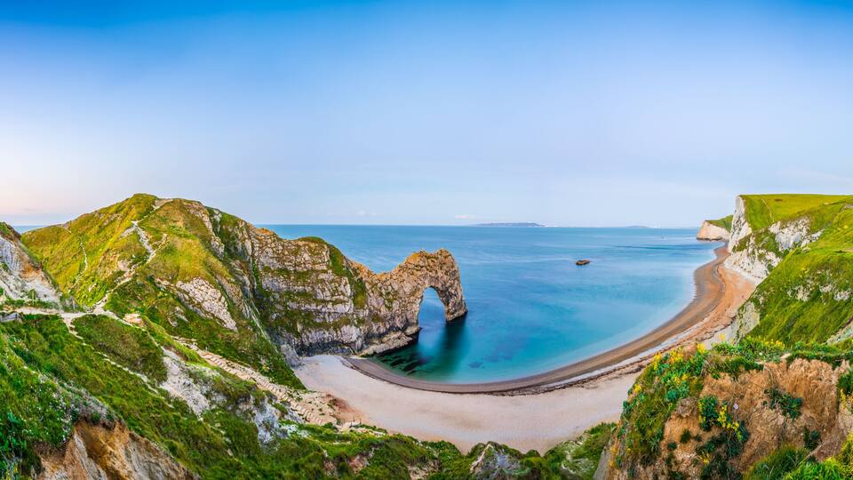 A coastal landscape with a prominent rock arch formation, a sandy beach, and a clear blue sea.