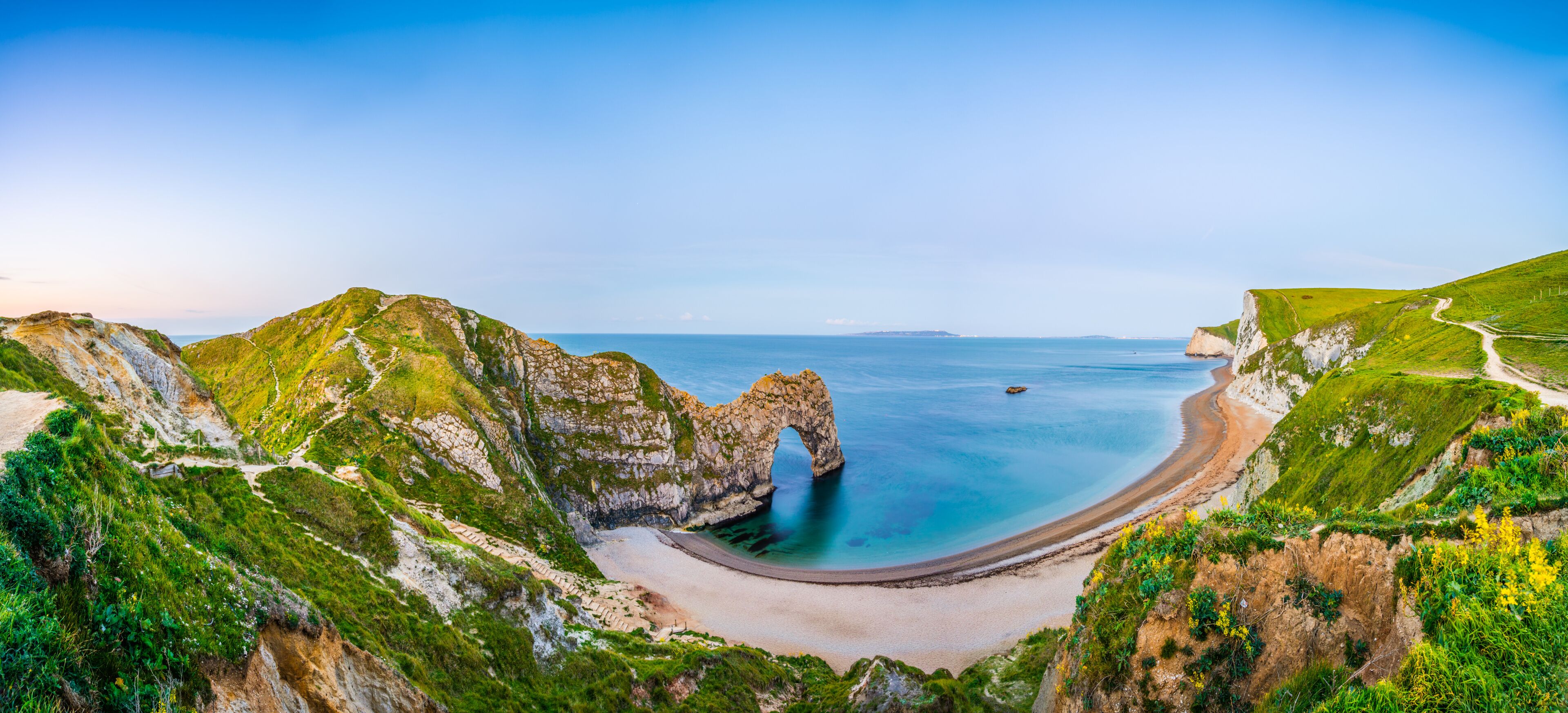 A coastal landscape with a prominent rock arch formation, a sandy beach, and a clear blue sea.