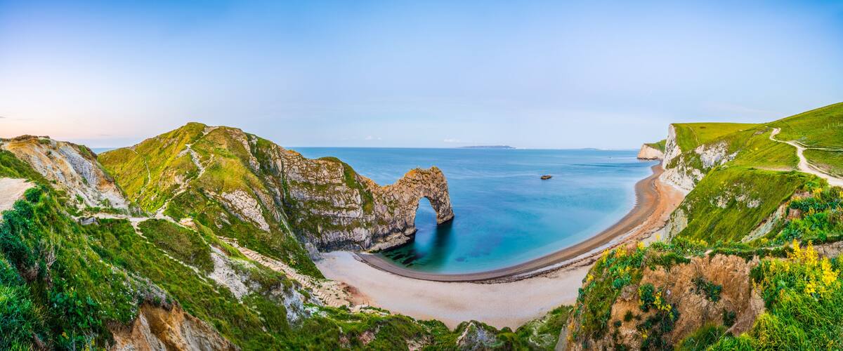 A coastal landscape with a prominent rock arch formation, a sandy beach, and a clear blue sea.