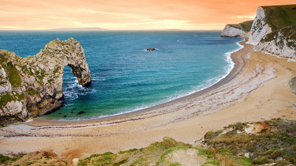 A coastal landscape with a prominent rock arch formation, a sandy beach, and cliffs.