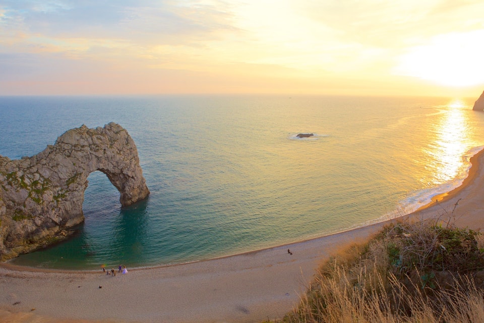 A coastal landscape with a natural rock arch formation, a sandy beach, and a clear view of the sea.