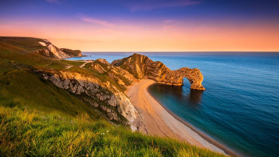 A coastal landscape with a prominent rock arch formation, a sandy beach, and a grassy hillside.