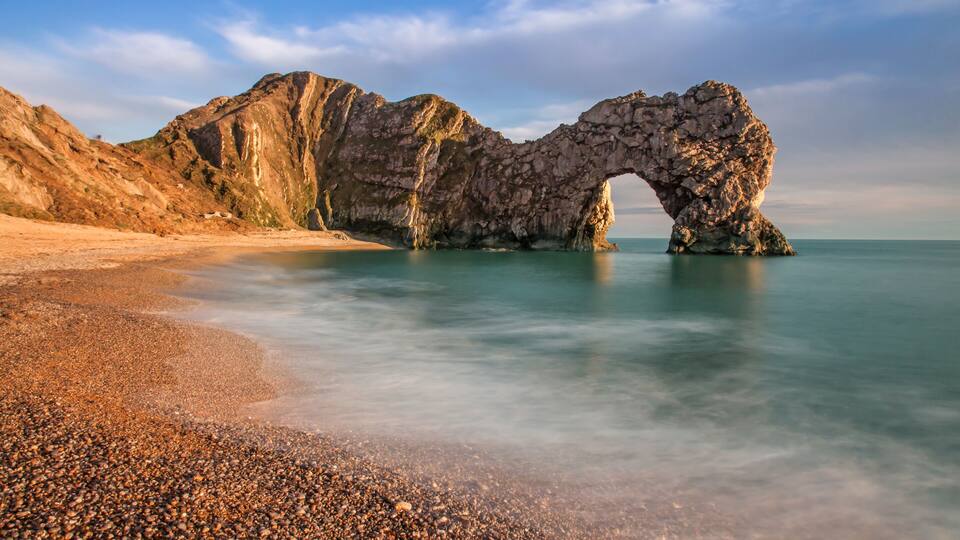 A natural rock arch formation over a beach with pebbles and calm water.