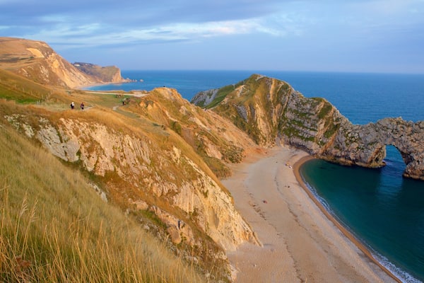 Eine Küstenlandschaft mit einer markanten Felsbogenformation, einem Sandstrand und sanften Hügeln.