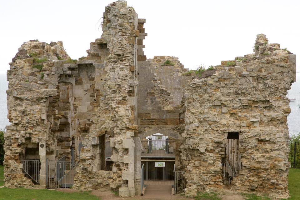 Restes de pedra amb una porta i un edifici petit al centre, amb vista a una massa d'aigua amb velers.