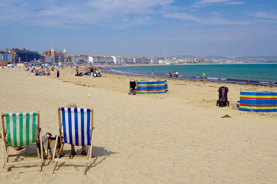 A beach with striped beach chairs and umbrellas, a sandy shore, and a cityscape in the background.