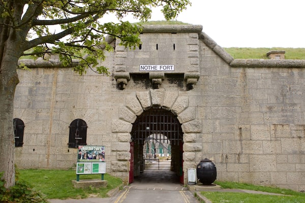 The entrance to Nothe Fort with a stone wall, a gate, and a signboard.