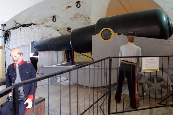 A large cannon in a museum exhibit, surrounded by metal railings and a display sign.