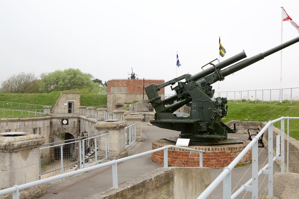 A large artillery piece in a historic fortification with flags and a small building in the background.