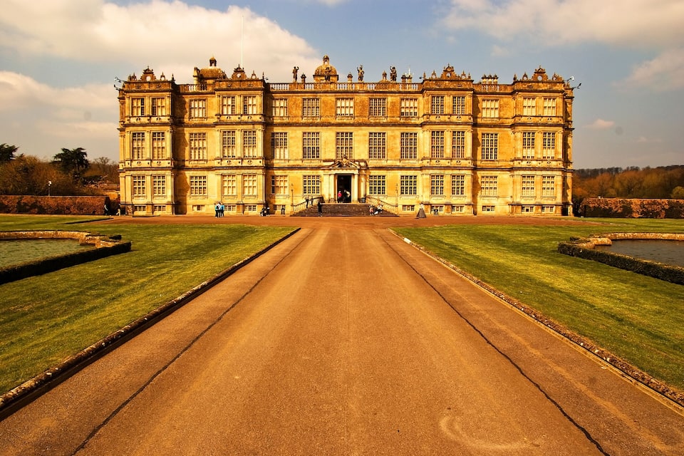 Um edifício grande e ornamentado, com fachada simétrica, entrada central e um jardim formal com lago.