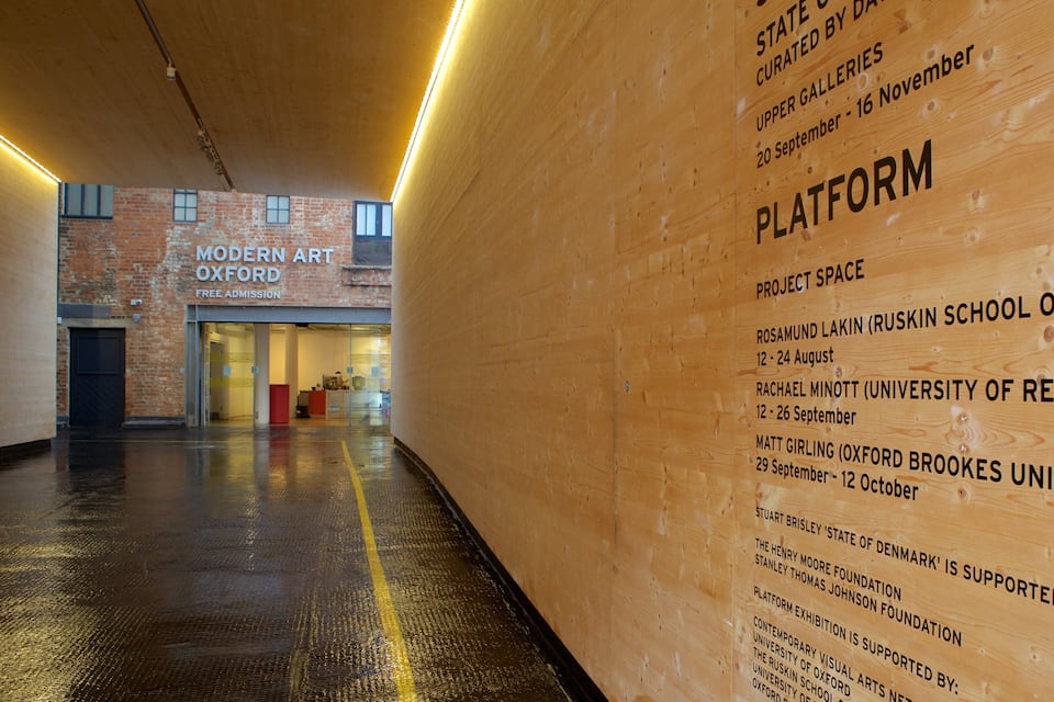 Een houten muur met tekst over een kunsttentoonstelling, een bakstenen gebouw met de tekst 'Modern Art Oxford', en een natte vloer.