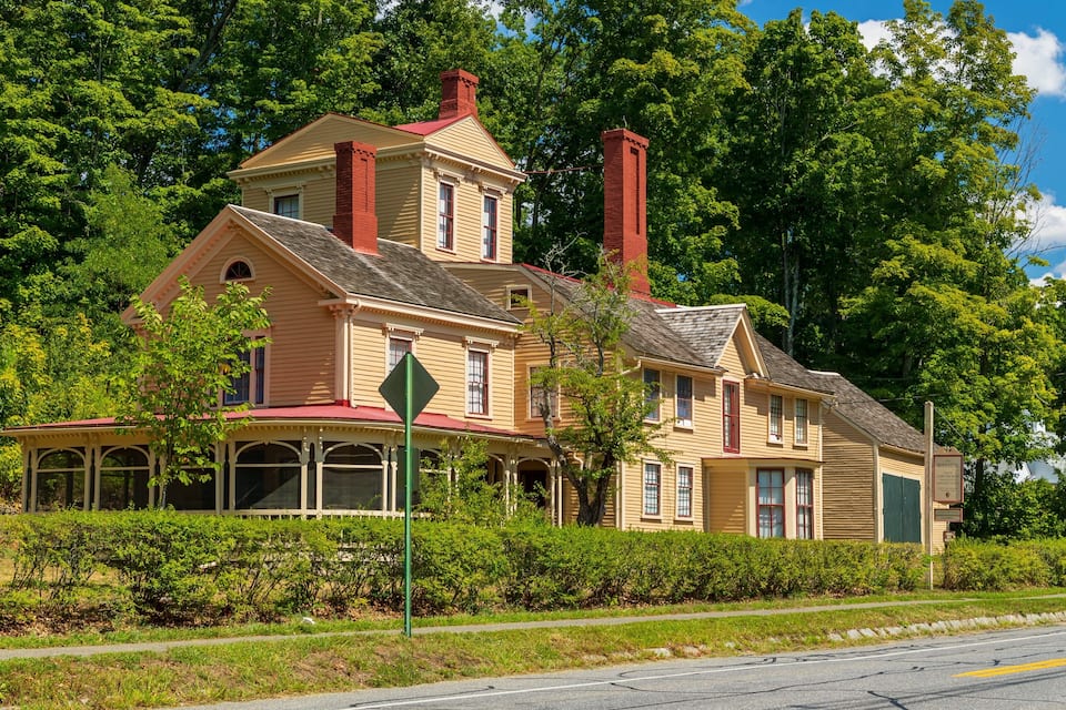Una casa grande y amarilla con techo rojo y chimeneas, rodeada de frondosos árboles verdes.
