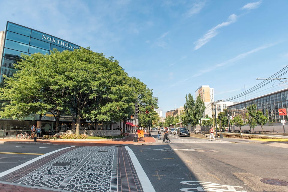 A city street with a large tree, a modern building labeled "Northeastern University," and a crosswalk.