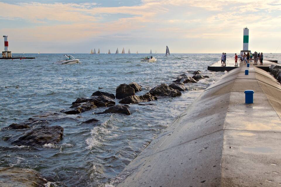 A pier with rocks in the water, a lighthouse, and sailboats in the distance.