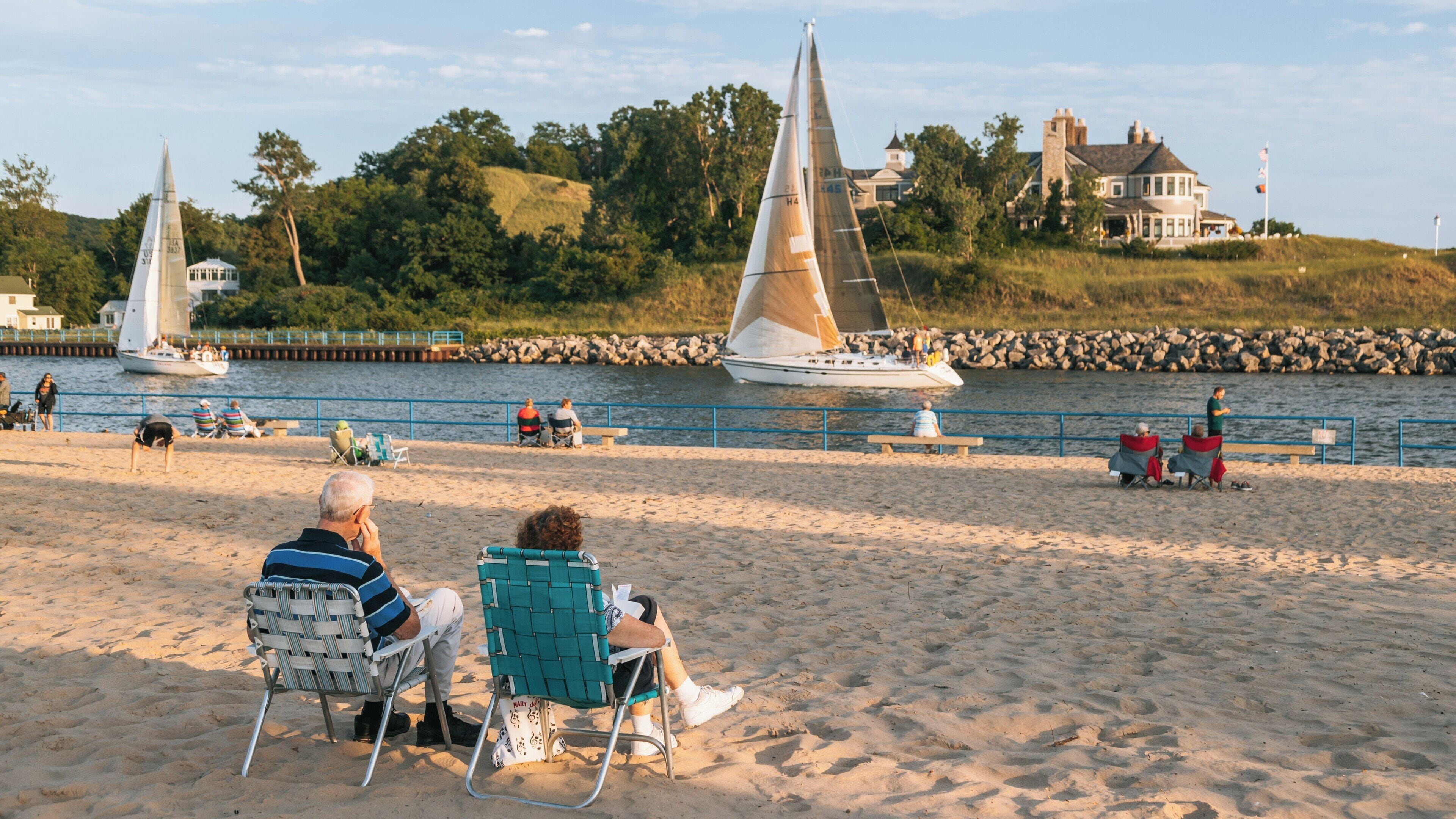 Scenic view at Holland State Park features beachgoers relaxing while sailboats glide across the water near Ottawa Beach in Michigan