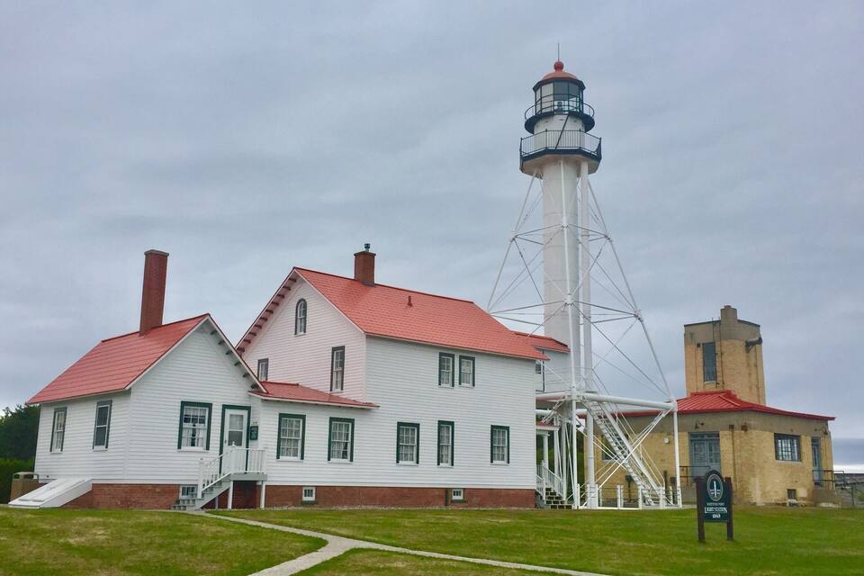 Un faro blanco con techo rojo y una torre de ladrillo junto a un edificio blanco con ventanas verdes.