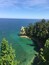 A coastal landscape with clear turquoise waters, a rocky cliff, and dense green foliage.