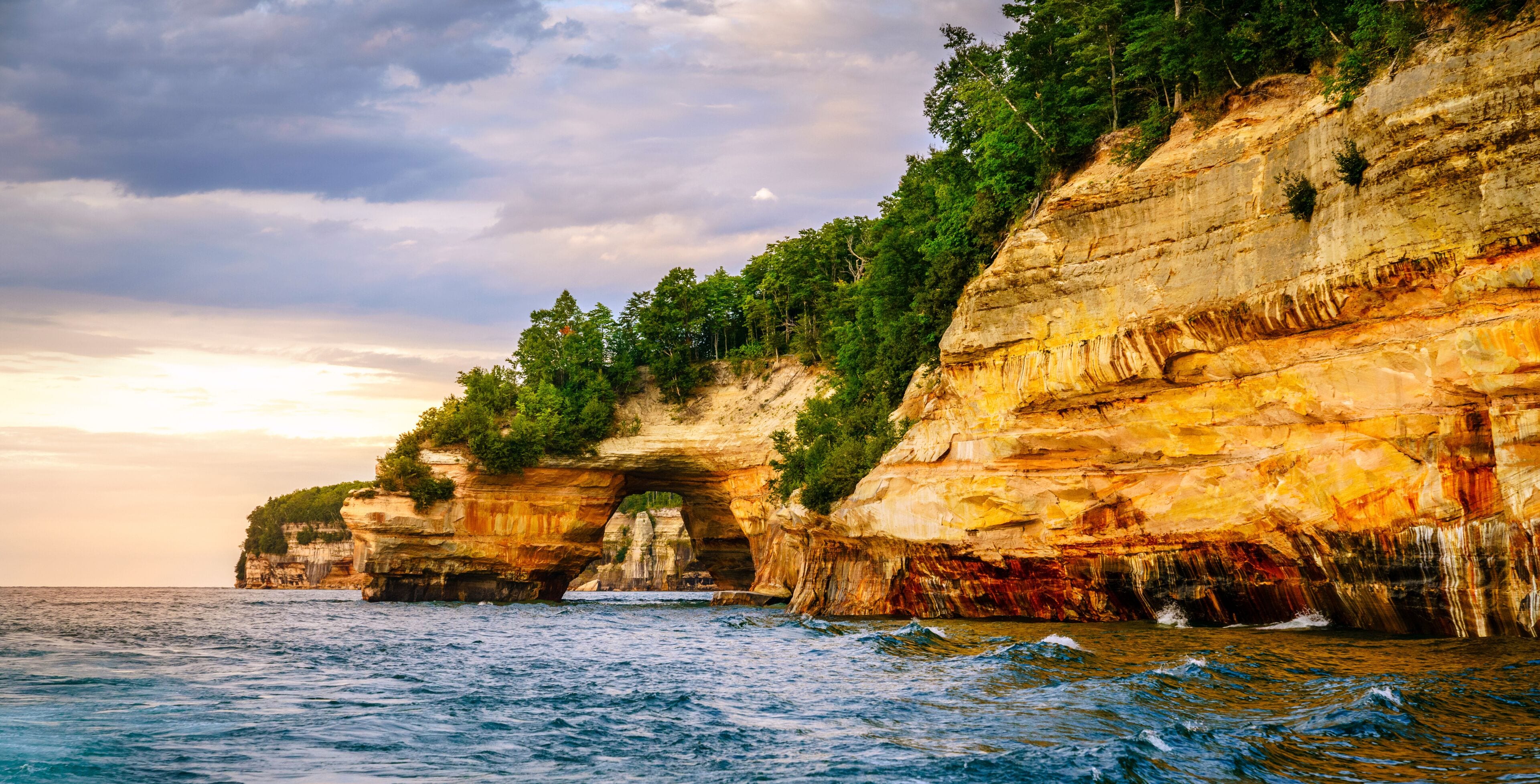 A coastal cliff with a natural arch formation, dense greenery, and a view of the sea.