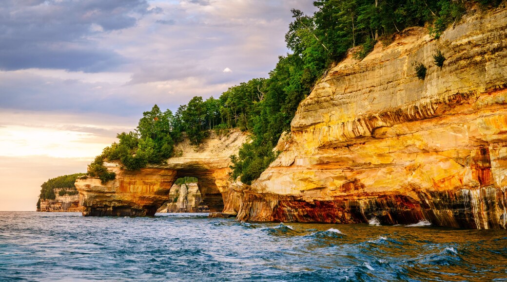 A coastal cliff with a natural arch formation, dense greenery, and a view of the sea.