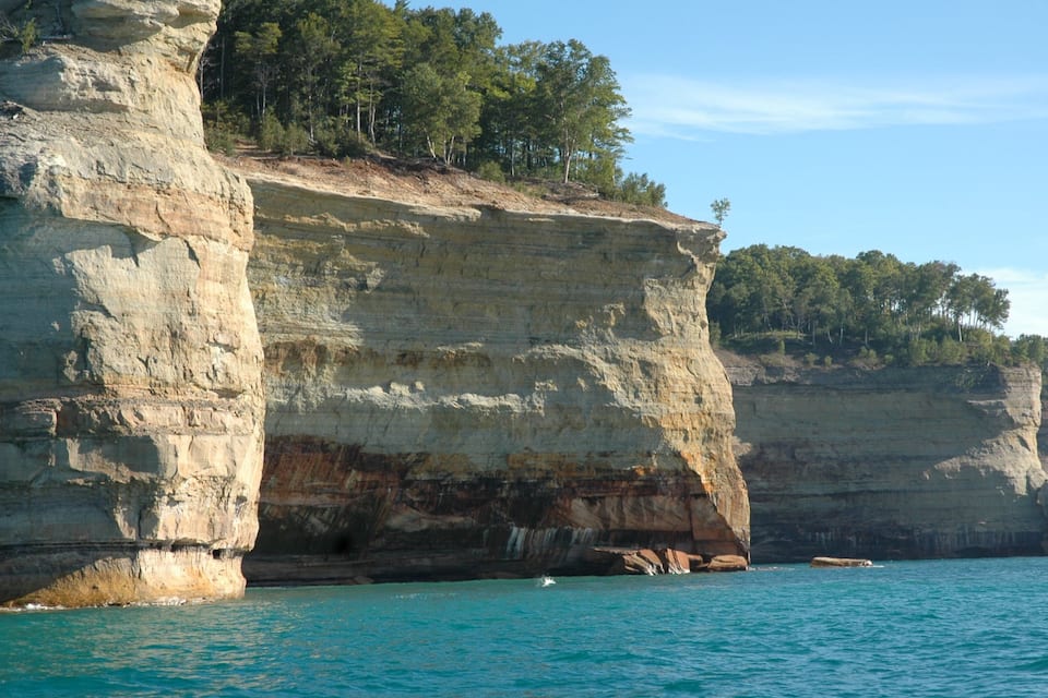 A coastal cliff with a clear blue sea and a forested hilltop.