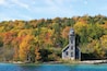 Un phare en bois se dresse au bord d’un lac aux eaux d’un bleu limpide, entouré d’arbres aux couleurs automnales.