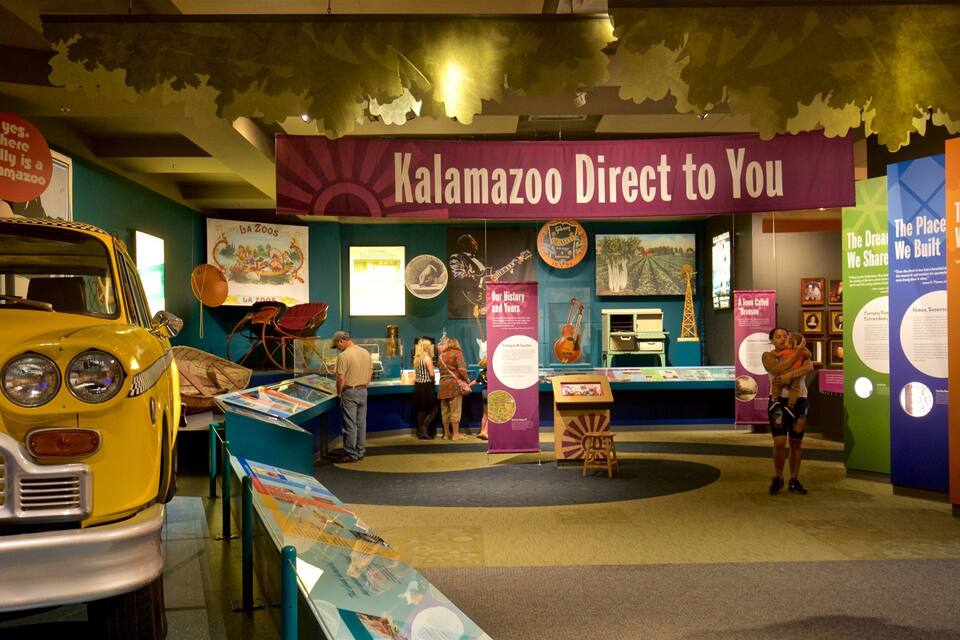Une exposition de musée comprenant une voiture jaune, une banderole violette sur laquelle on peut lire « Kalamazoo, directement chez vous », et divers panneaux d’information.