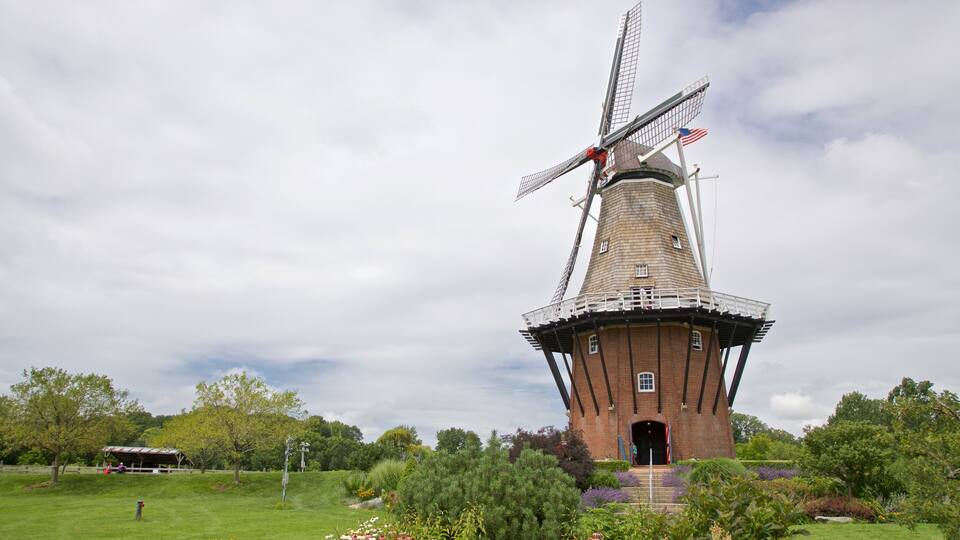 Un molino de viento con una bandera en la parte superior, rodeado de un área verde con árboles y un edificio pequeño al fondo.