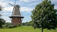 Eine traditionelle Windmühle mit Backsteinfundament und weißer Windhebel, umgeben von üppigem Grün und wolkenlosem Himmel.