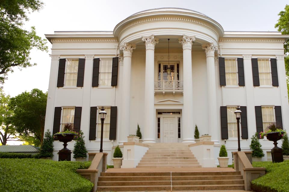 A grand white building with black shutters, a central balcony, and a symmetrical facade.