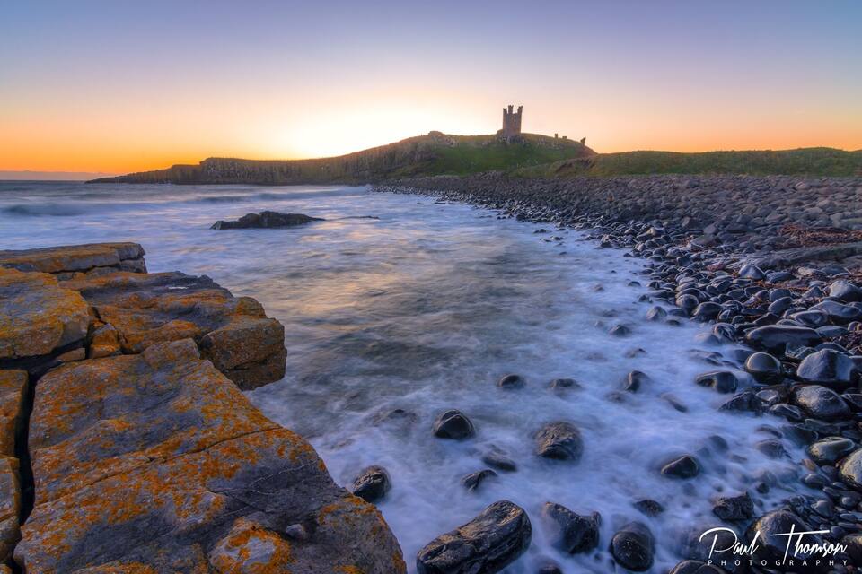 A coastal landscape with a castle on a hill, rocky shoreline, and ocean waves.